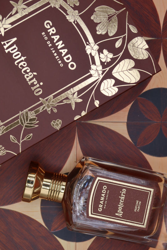 Granado Apotecário perfume bottle with gold cap beside its burgundy and gold illustrated box on patterned marquetry tiles, notes of bergamot, cardamom, black pepper, leather, black tea, myrrh and tonka