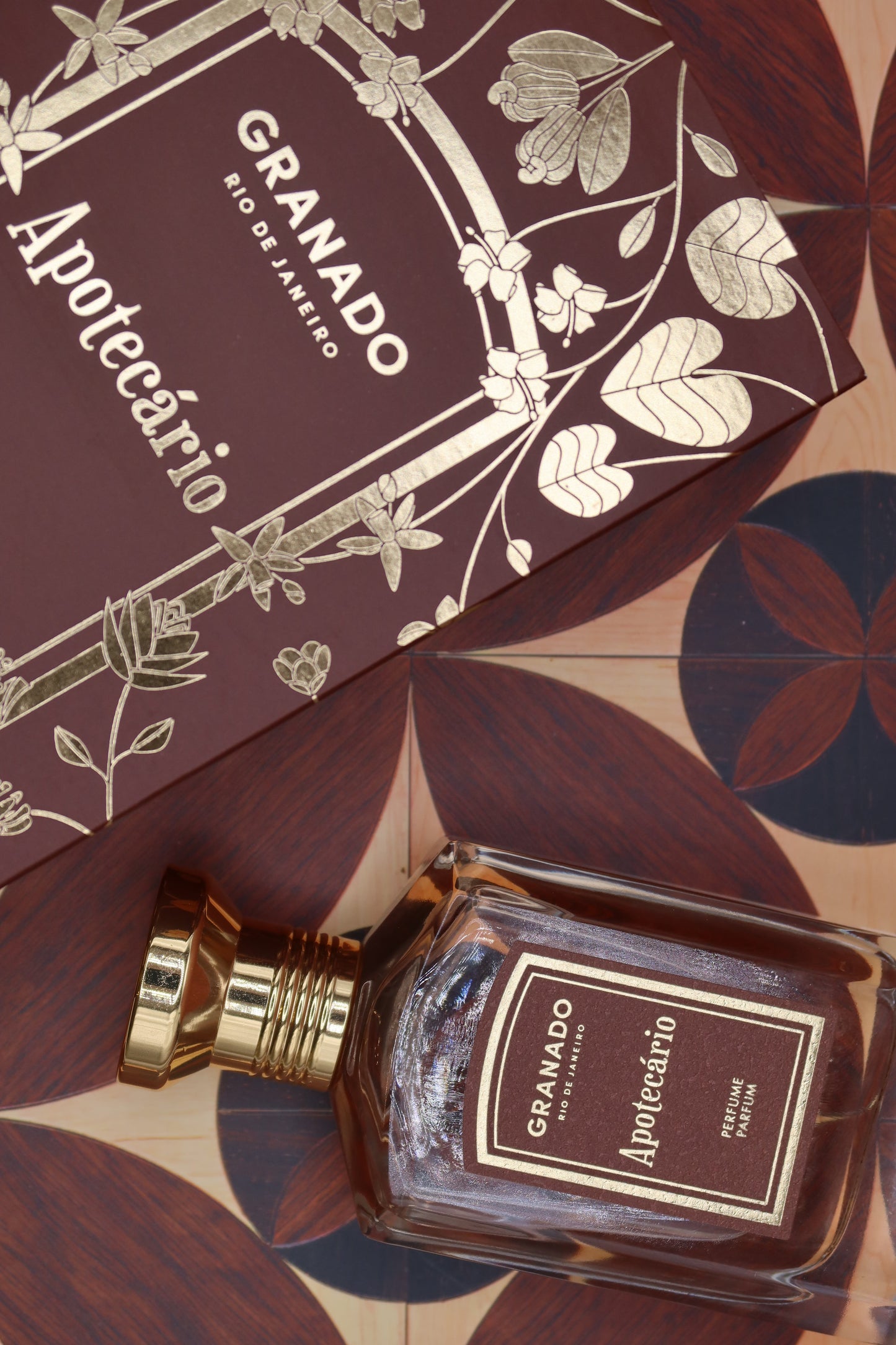 Granado Apotecário perfume bottle with gold cap beside its burgundy and gold illustrated box on patterned marquetry tiles, notes of bergamot, cardamom, black pepper, leather, black tea, myrrh and tonka