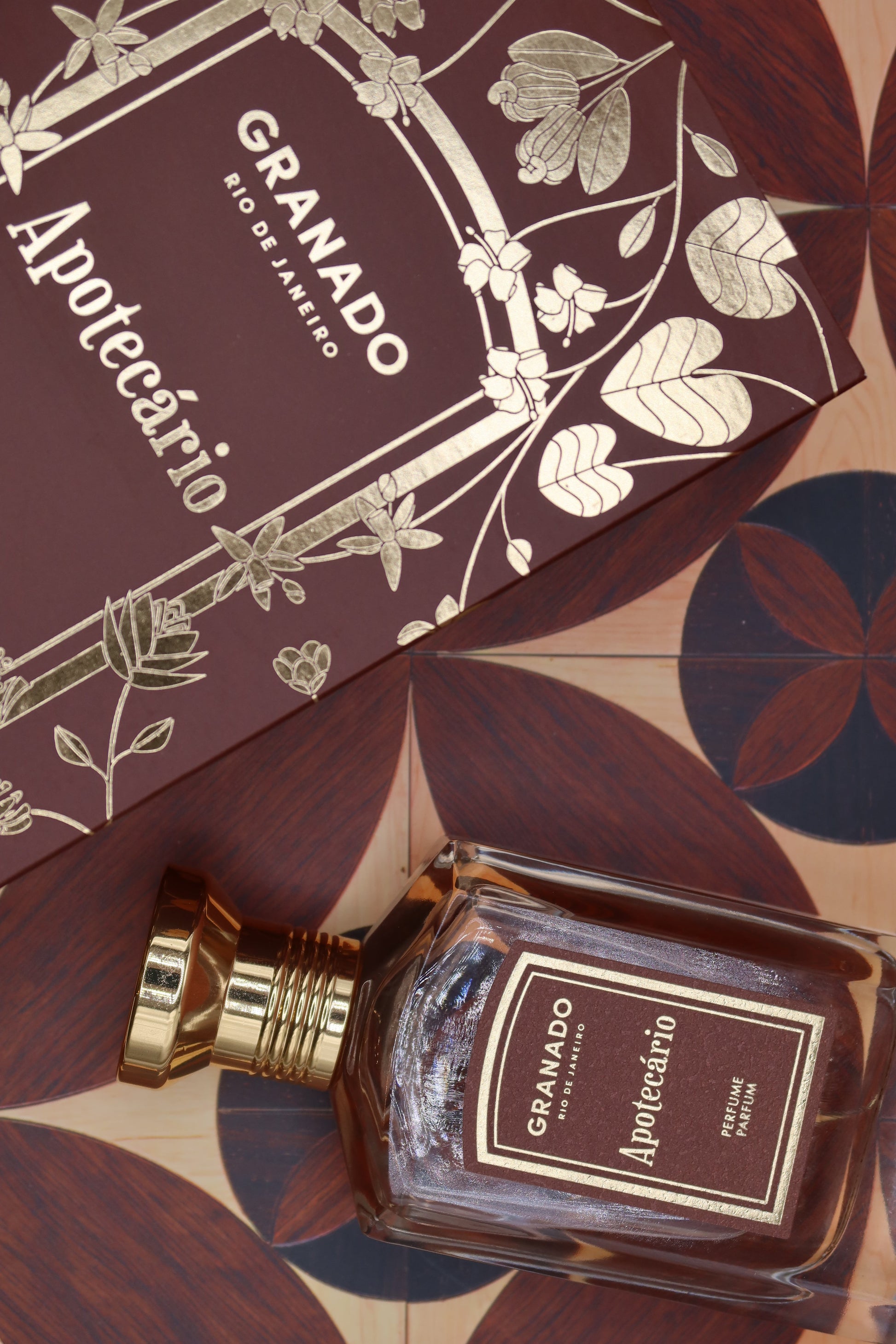 Granado Apotecário perfume bottle with gold cap beside its burgundy and gold illustrated box on patterned marquetry tiles, notes of bergamot, cardamom, black pepper, leather, black tea, myrrh and tonka